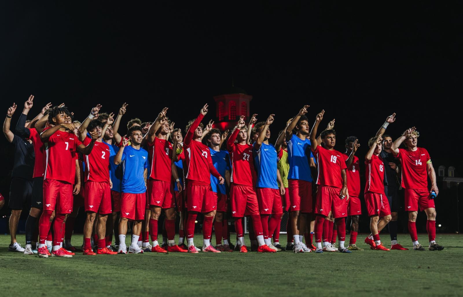 SMU men's soccer players in red kits celebrate together on the field at night (photo: SMU Athletics)