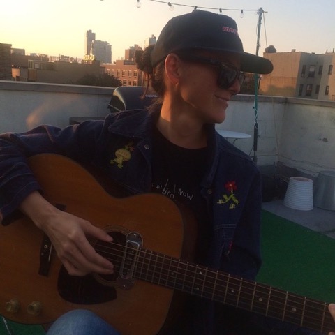 Ambassador Parsley (Ambrosia Parsley) smiling on a rooftop at sunset, wearing a cap and sunglasses and holding an acoustic guitar.