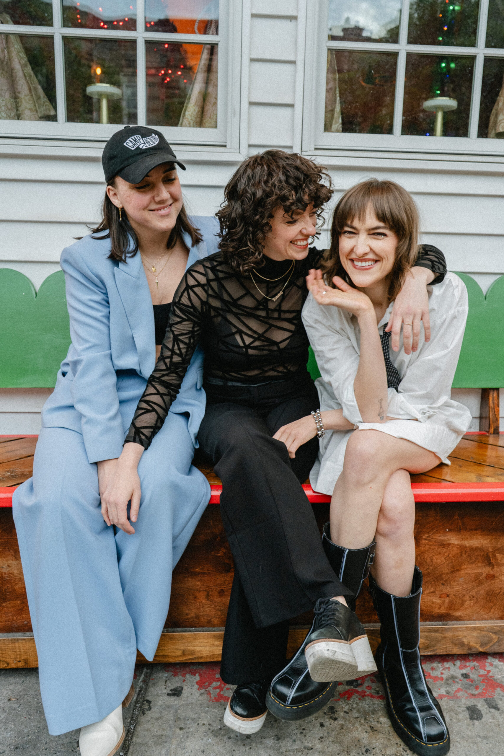Brooklyn folk-rock trio Camp Bedford, Roxanne Quilty, Kailey Prior and Rebekah Farr, sitting together laughing on a bench in a photo by Brooke Banister