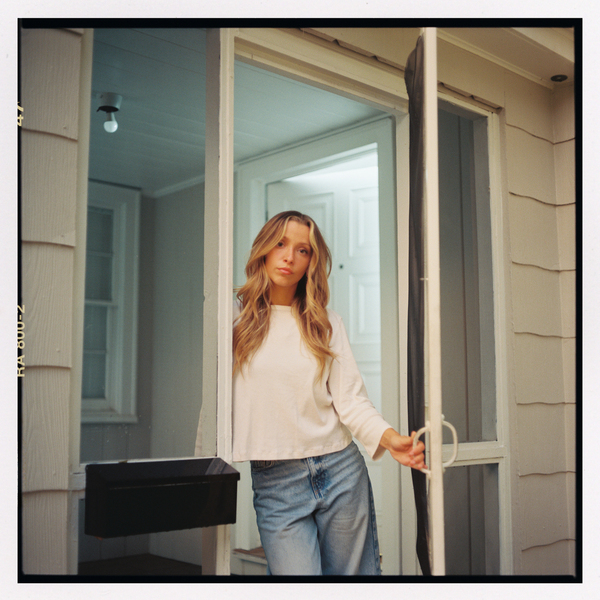 Chelsea Plank standing in a doorway holding a screen door open, wearing a white sweater and jeans
