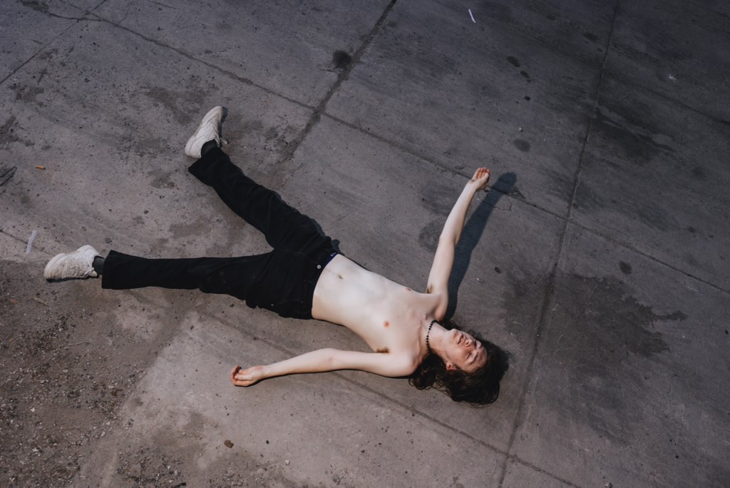Heathers (Heath Michael Thomas) lying on his back on concrete with his arms spread, photographed from above
