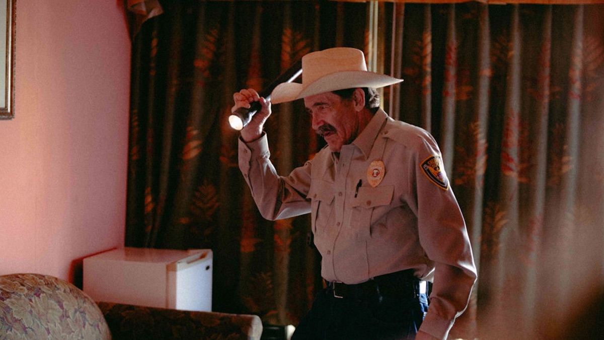 A uniformed police officer in a cowboy hat searches a motel room with a flashlight in a still from Jason Scott & The High Heat's "How to Get Away with Murder" music video