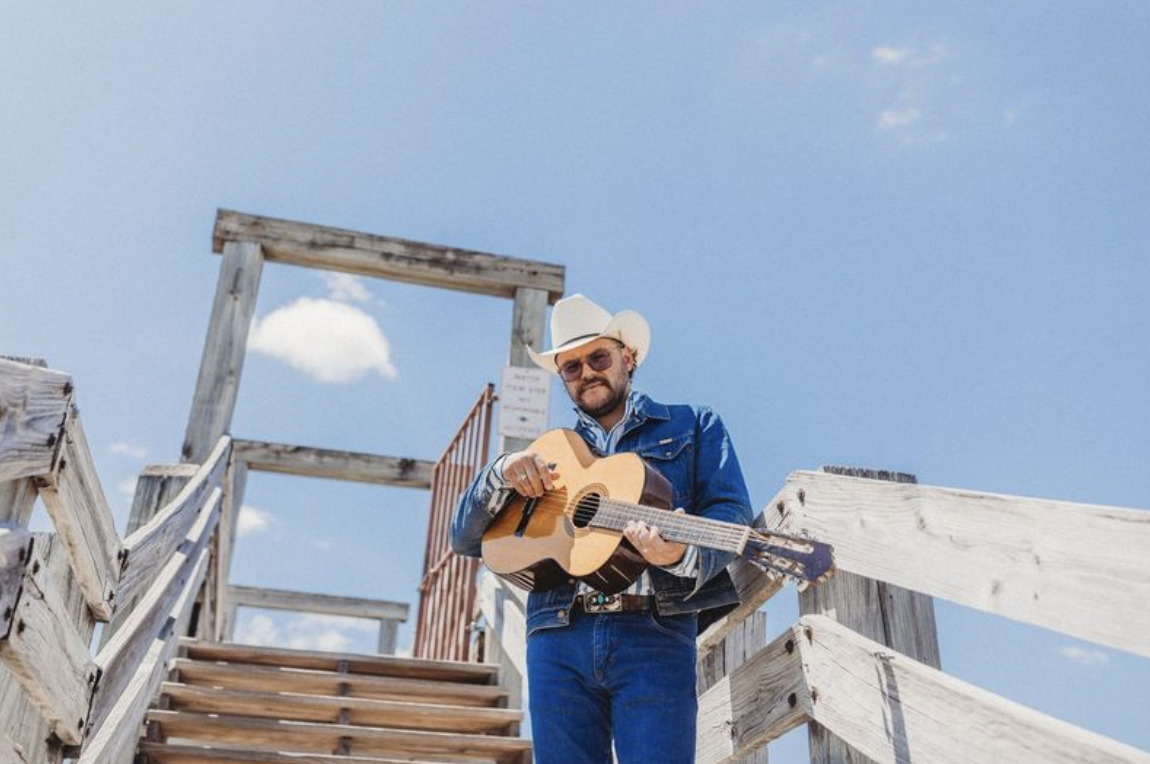 Josh Weathers in a white cowboy hat and denim jacket holding an acoustic guitar on wooden outdoor stairs under a blue sky
