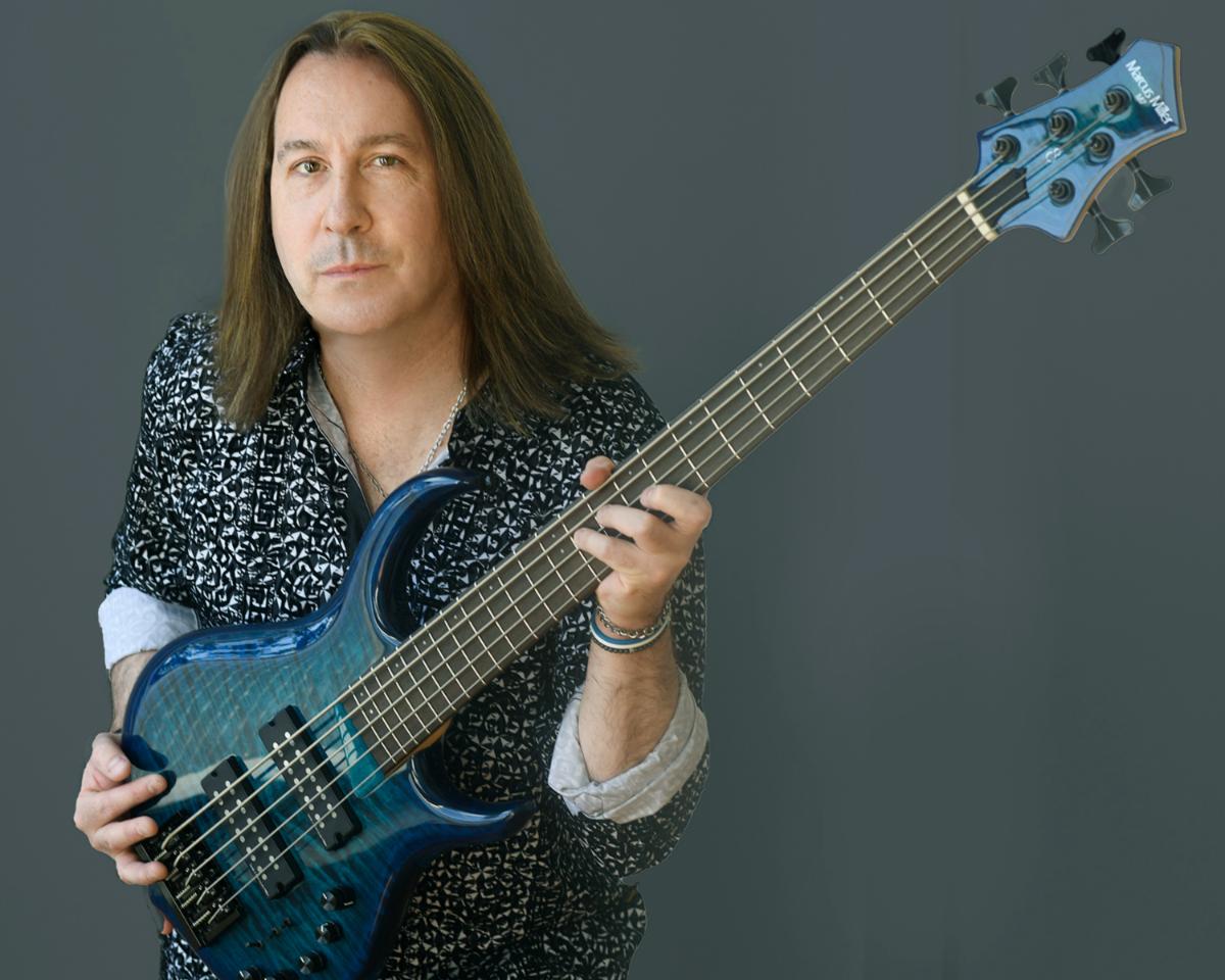Thomas Miller holding a blue five-string bass guitar against a grey backdrop.