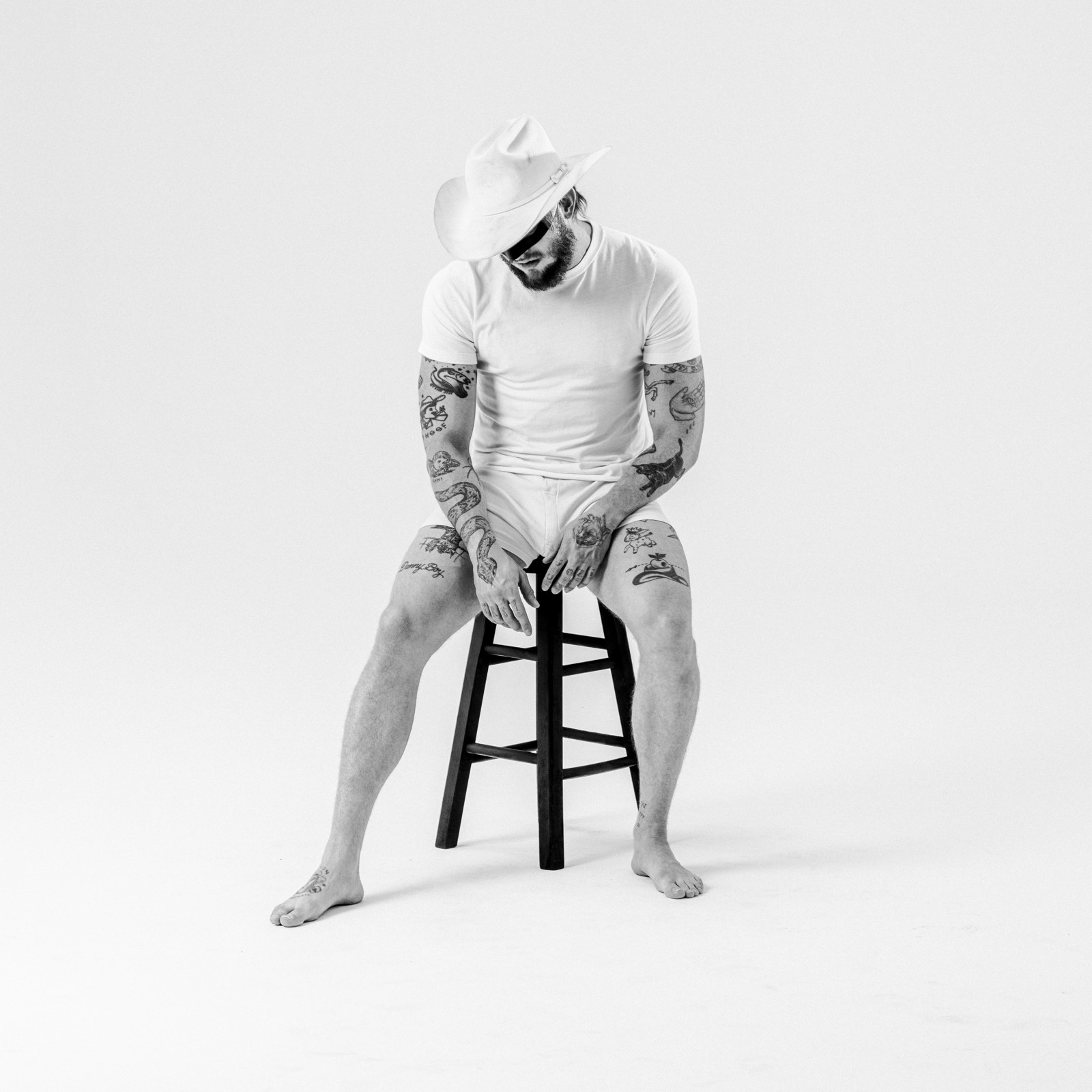 Orville Peck in a black-and-white press photo, masked and wearing a white cowboy hat and white t-shirt, his tattooed arms resting on a stool (Warner Records)