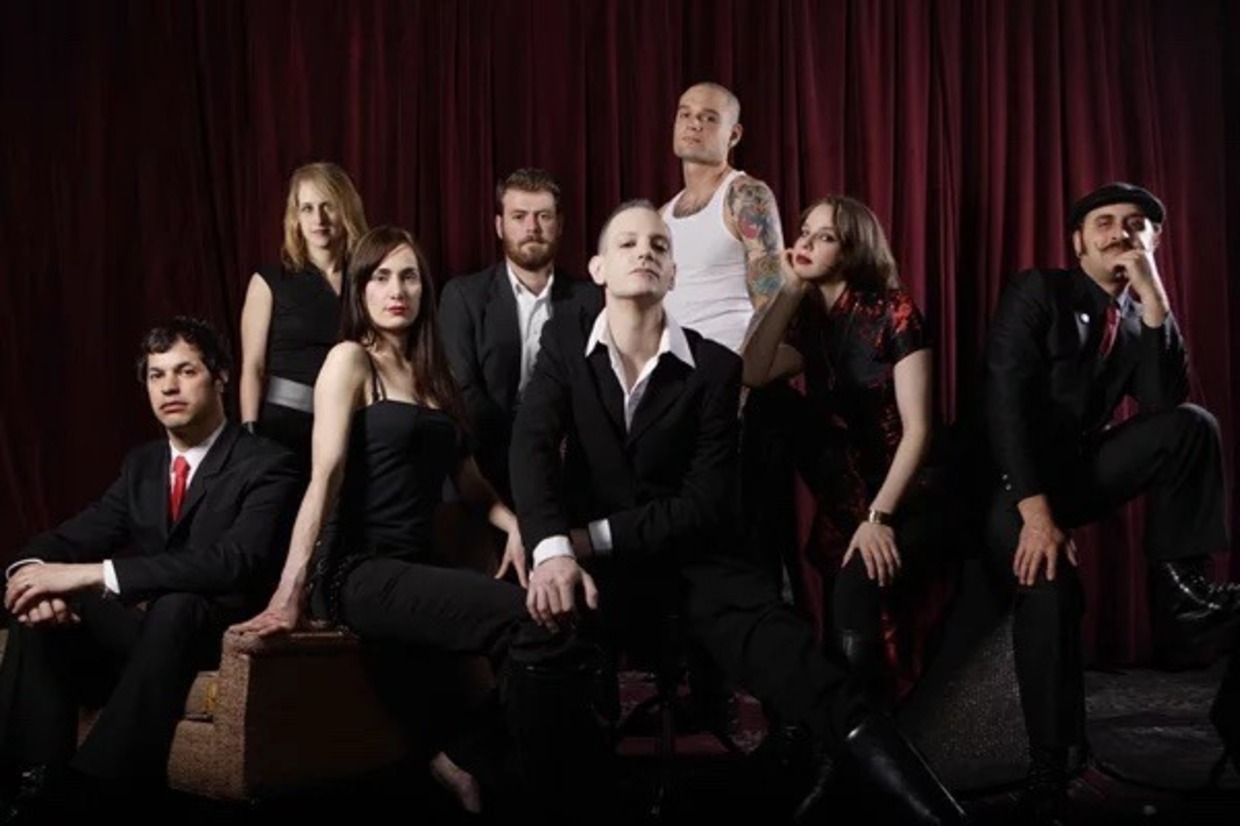 The World/Inferno Friendship Society posed in black formal wear against a red stage curtain, with singer Jack Terricloth seated in the center