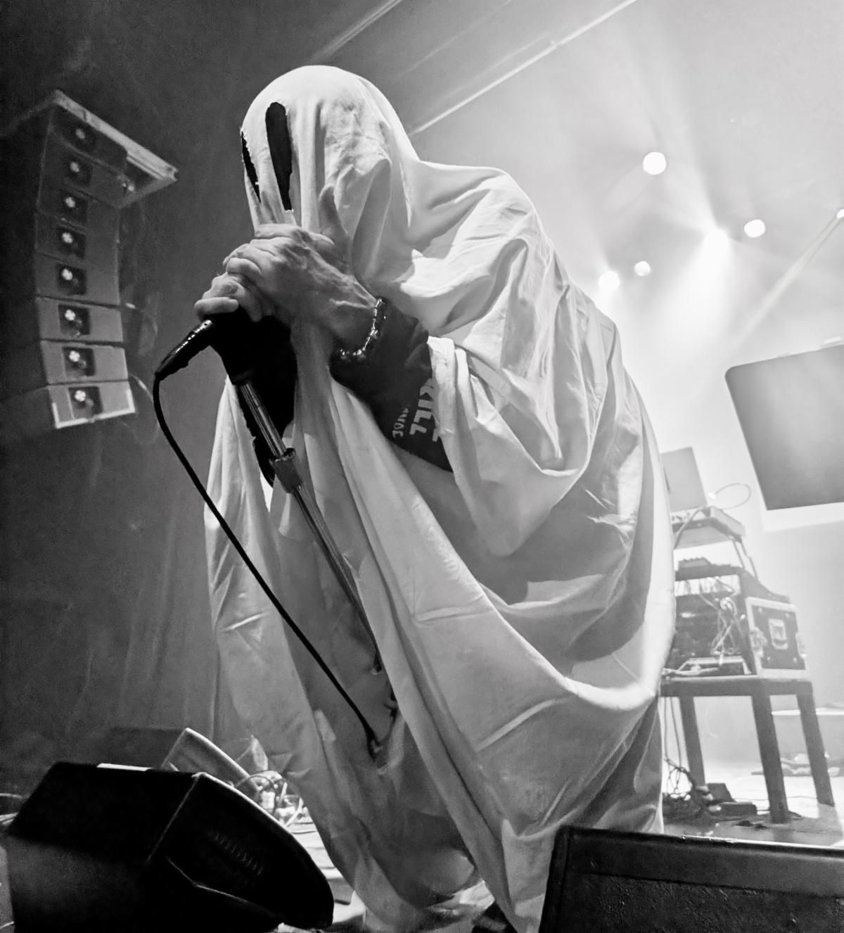 MALHAVOC frontman Jimi LaMort performing live in a white ghost costume, gripping a microphone on stage — photo by Natasha Rego de Assunçao