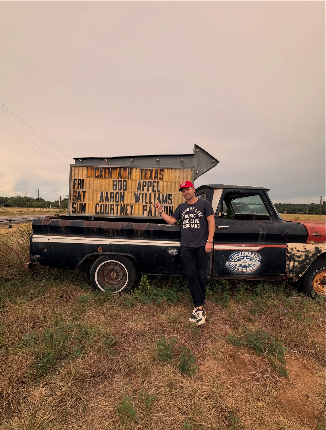 Aaron Williams standing beside a vintage pickup truck under the Luckenbach, Texas marquee sign listing his name on the bill