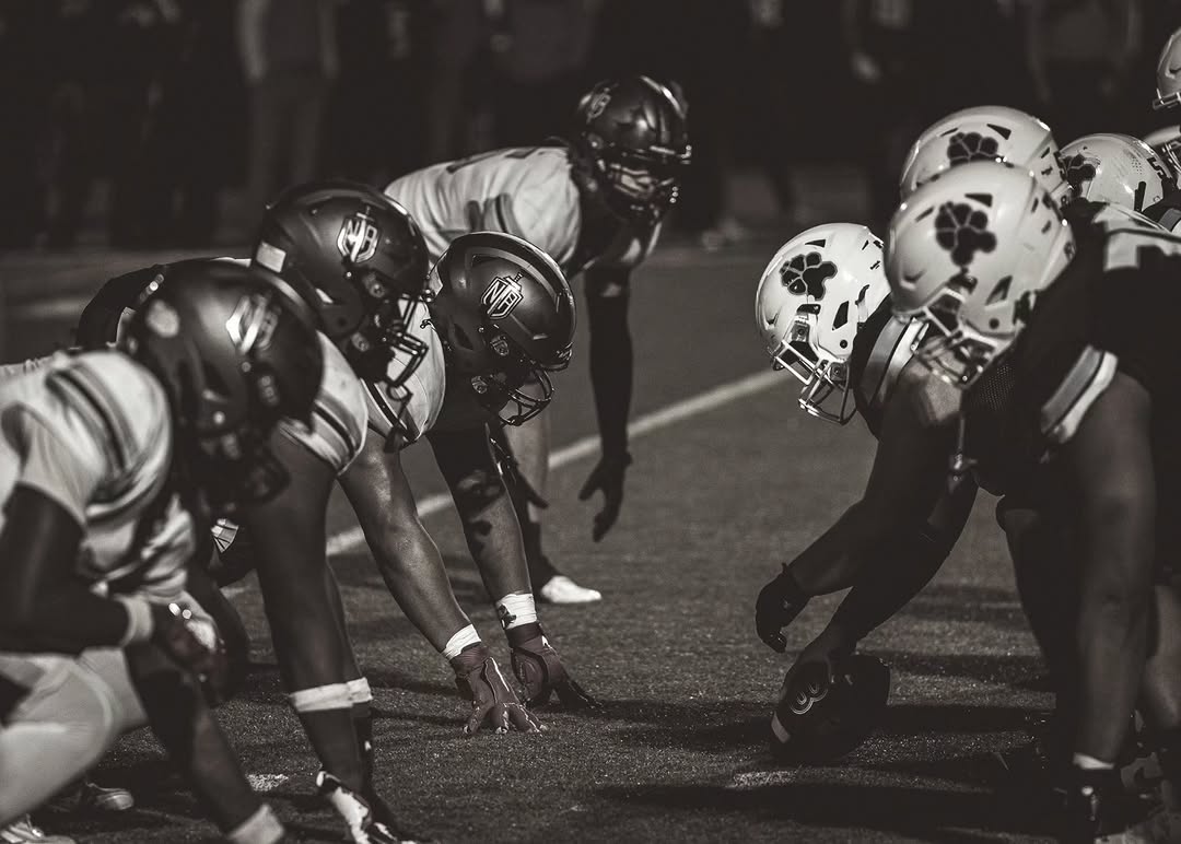 In a black and white photo, the North Atlanta offensive line, wearing dark NA helmets, sets at the line of scrimmage across from the defense during a night game