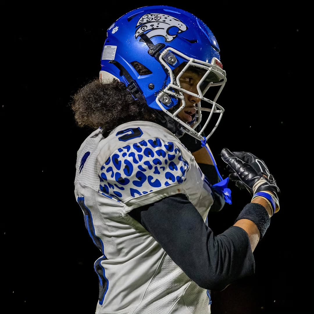 A Stephenson Jaguars player in a white jersey with blue jaguar-print shoulders and a blue jaguar helmet adjusts his gloves during a night game