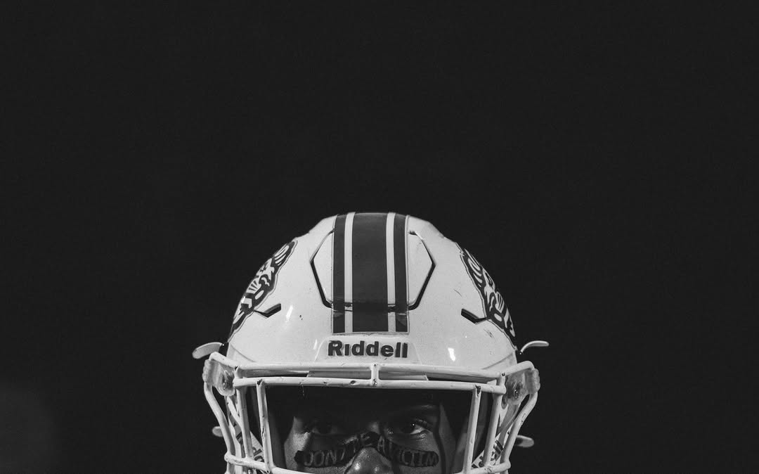 Black and white close-up photograph of a Peachtree Ridge player wearing a white helmet with the Lions mark and a center stripe, looking through his facemask at night