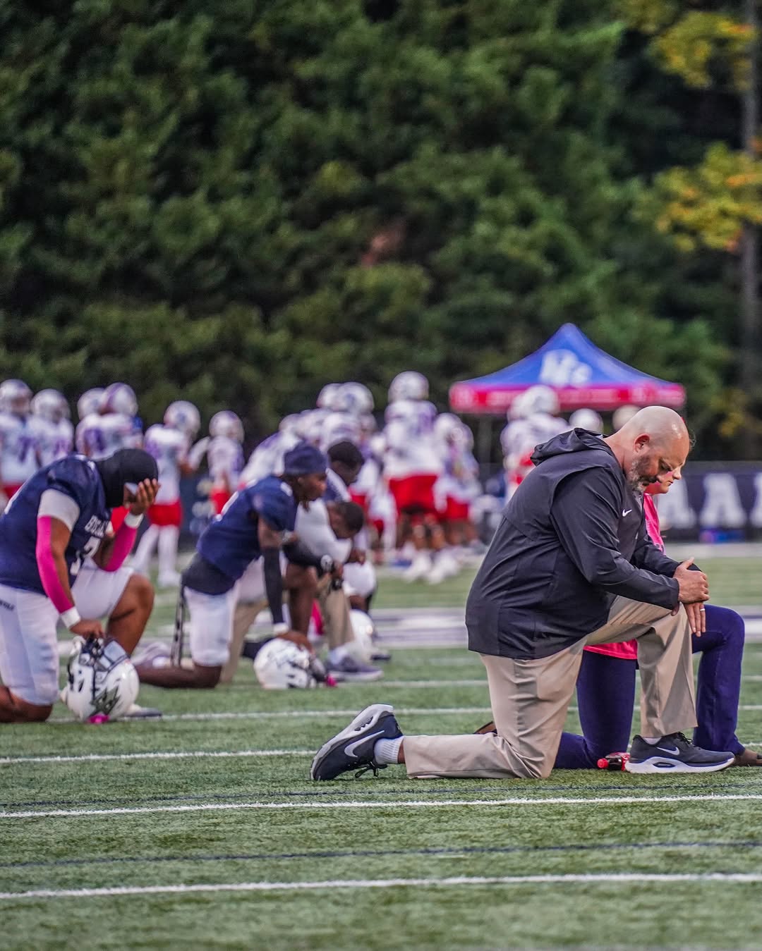 Norcross players in navy jerseys and their coach kneel on the field in a quiet moment before a game, helmets on the turf beside them