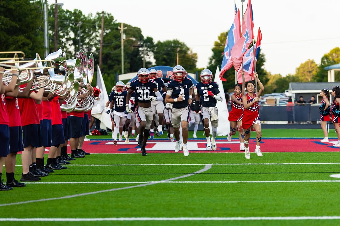 The Berkmar Patriots run onto their home field behind flag carriers and cheerleaders, with the marching band lining the entrance, numbers 30, 4 and 2 leading the team out
