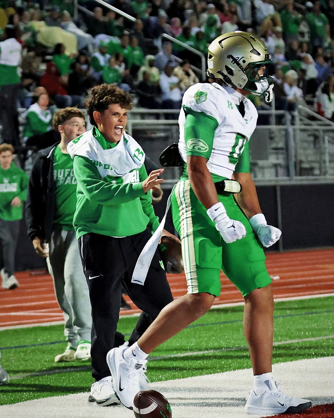 A Buford player in a white jersey, green pants and gold helmet celebrates on the field beside a teammate in team gear, with the home crowd in the stands behind them