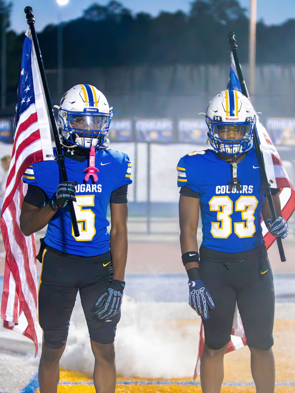 Two Chattahoochee Cougars players in blue jerseys, numbers 5 and 33, carry American flags as they lead the team onto the field at night