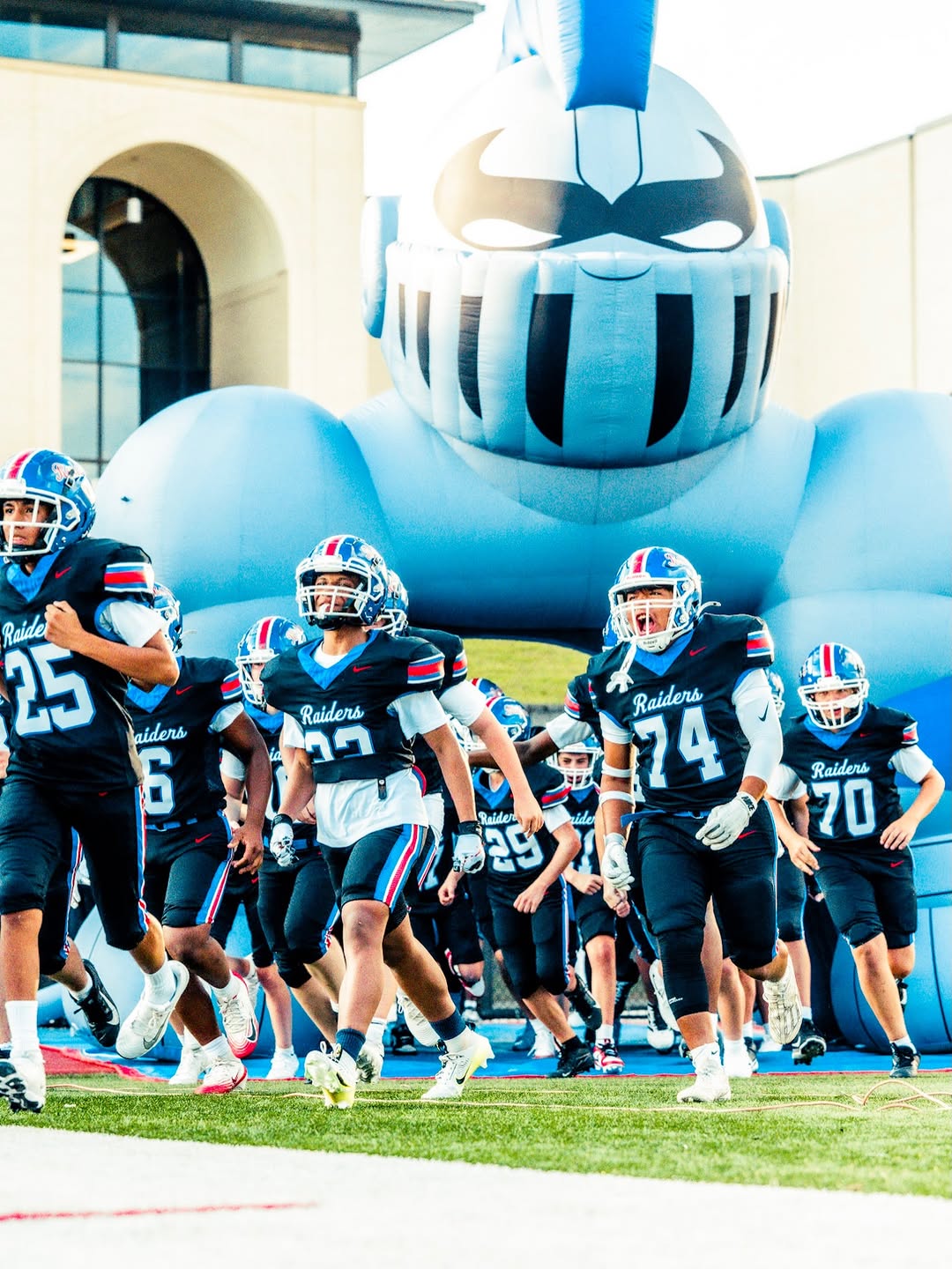 Riverwood Raiders players in black jerseys and blue helmets run onto the field through the team's inflatable Raider helmet tunnel