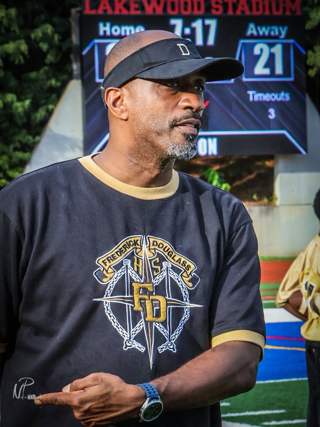 A Frederick Douglass coach in a black and gold Astros shirt and visor stands on the sideline at Lakewood Stadium with the scoreboard behind him during a game
