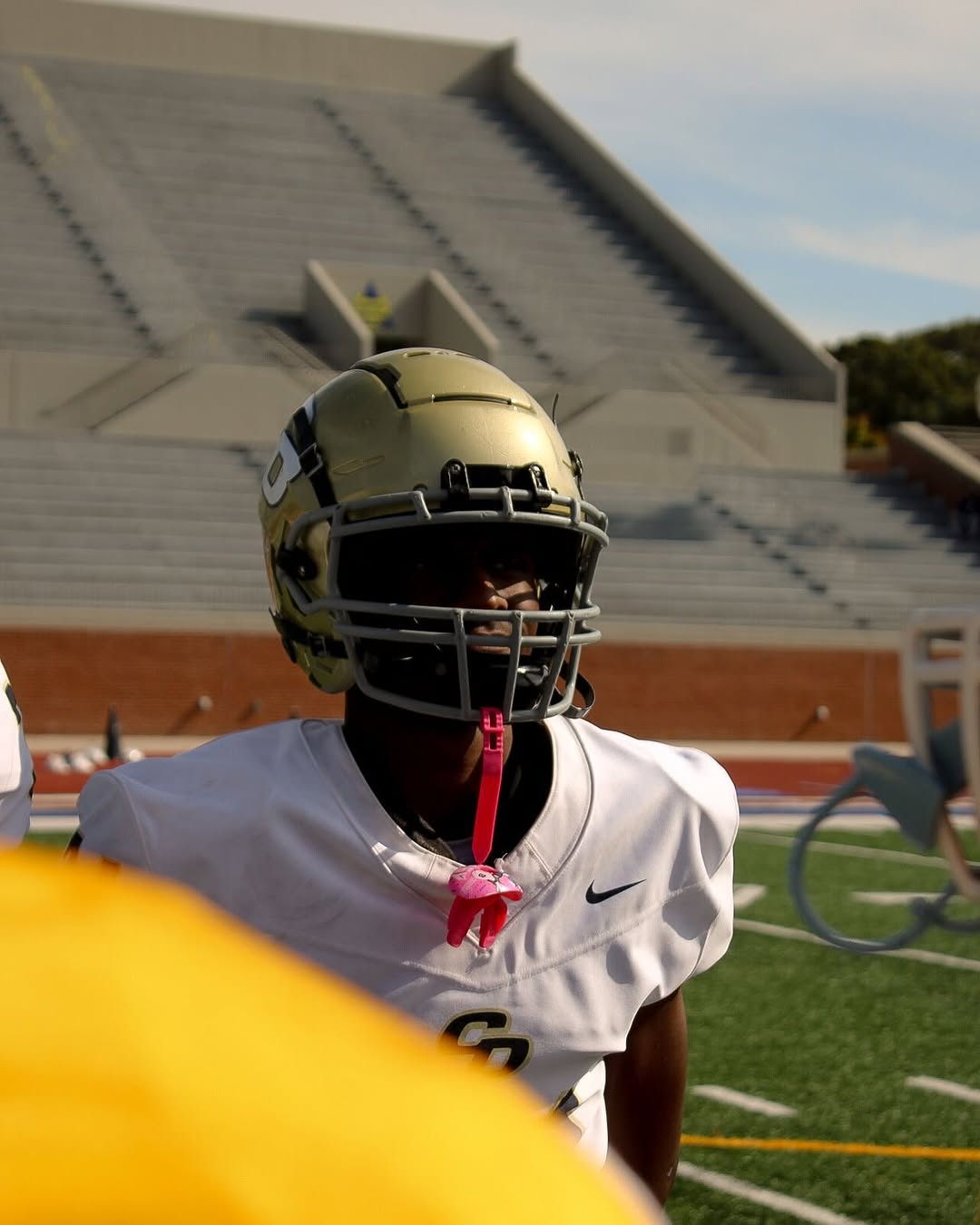 A Charles R. Drew Titans player in a white jersey and gold CD helmet stands on the field in front of the stadium bleachers on game day