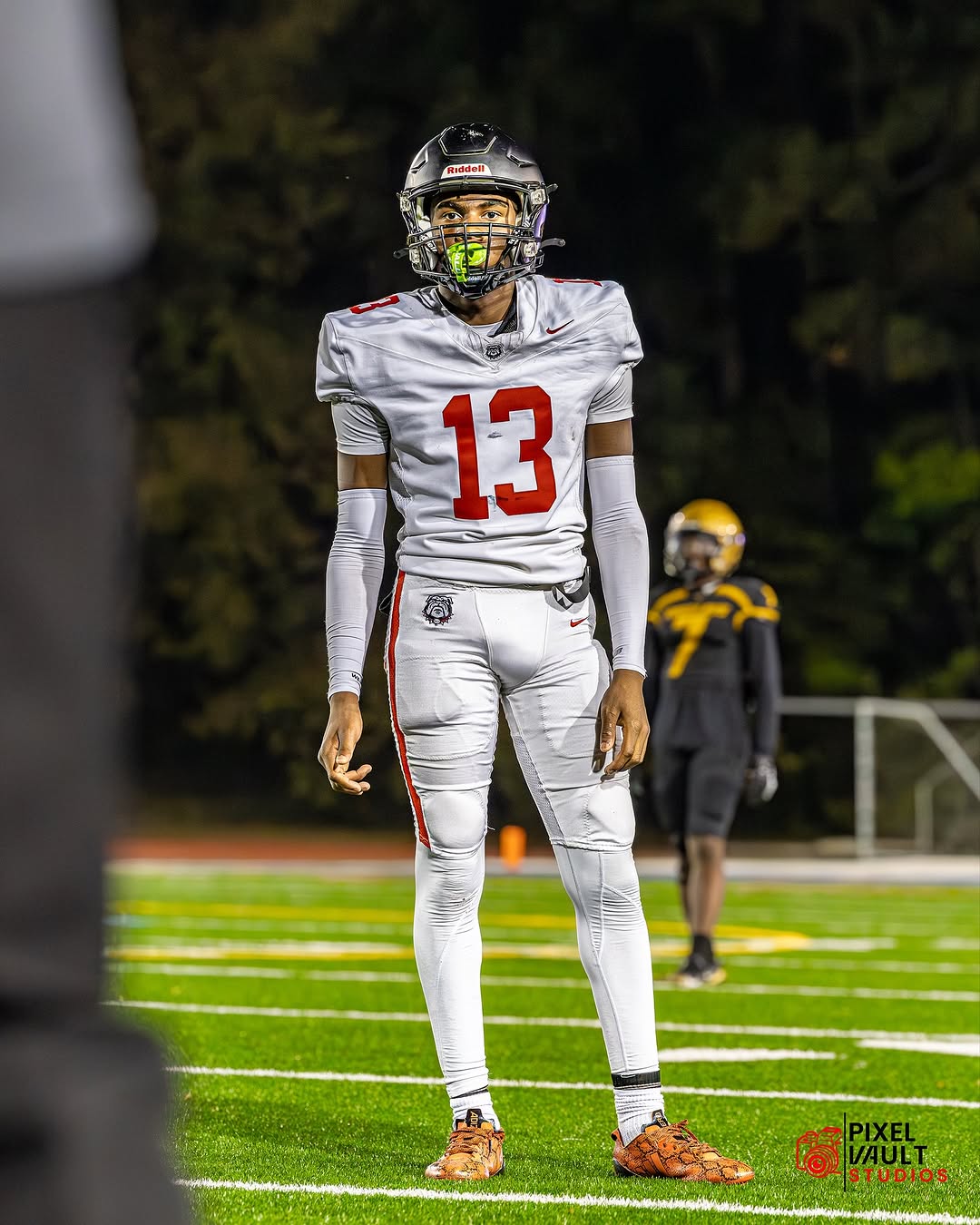 A Mt. Zion Bulldogs player wearing a white jersey with red No. 13 and a black helmet stands on the field during a night game