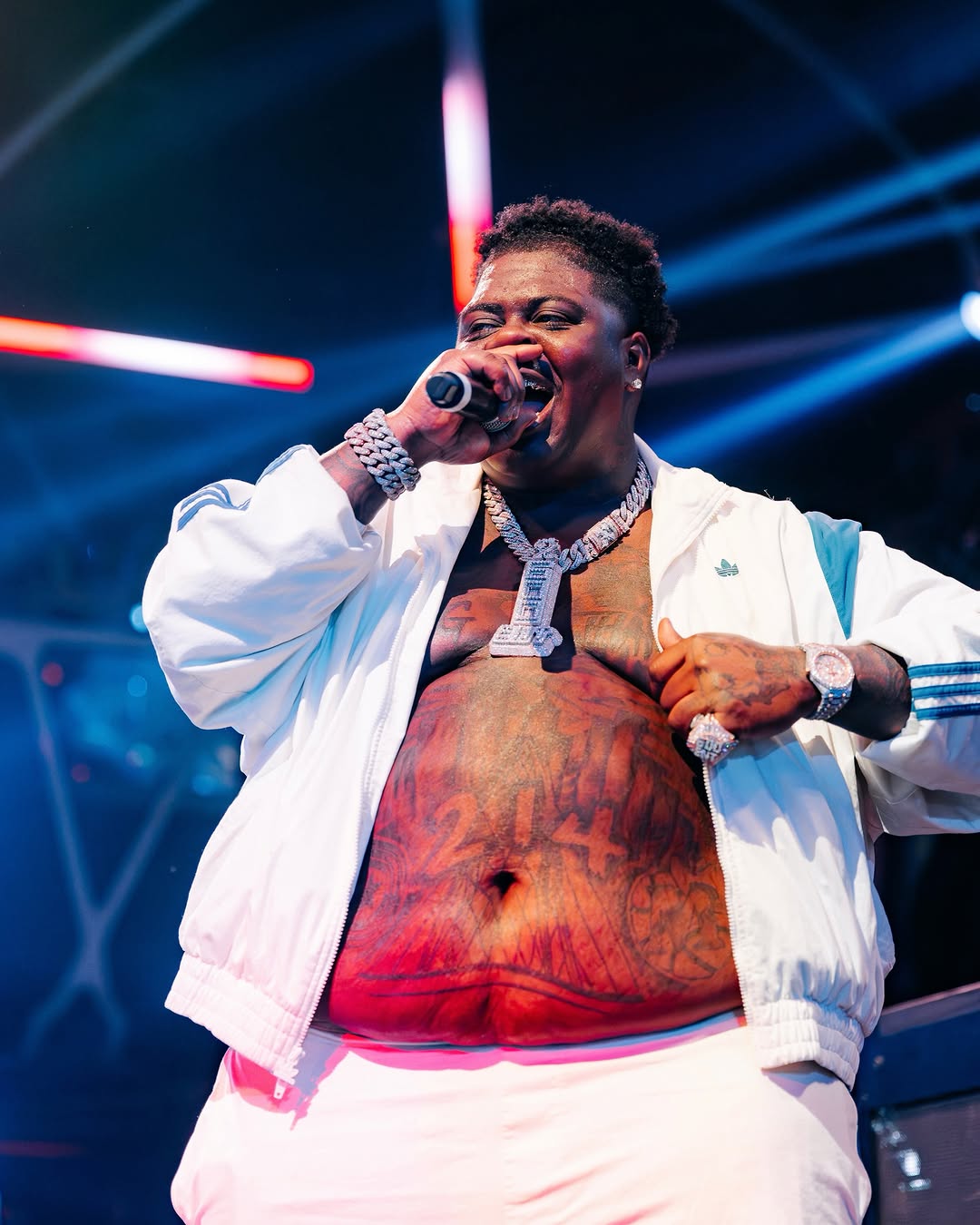BigXthaPlug performs on stage in a white Adidas jacket, holding a microphone, with his 214 chest tattoo showing.