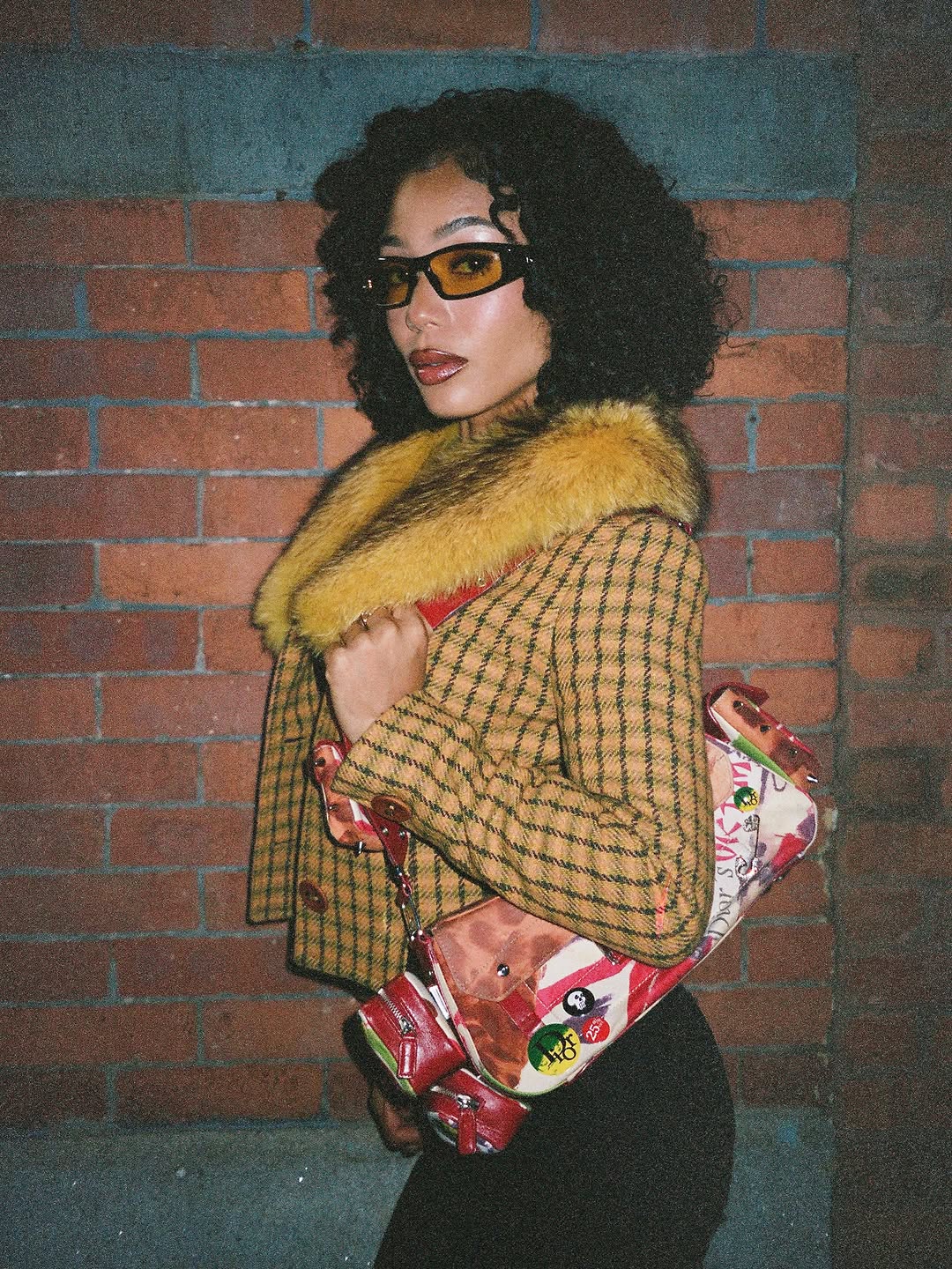 Coi Leray poses against a brick wall in a plaid jacket with a fur collar and tinted sunglasses.