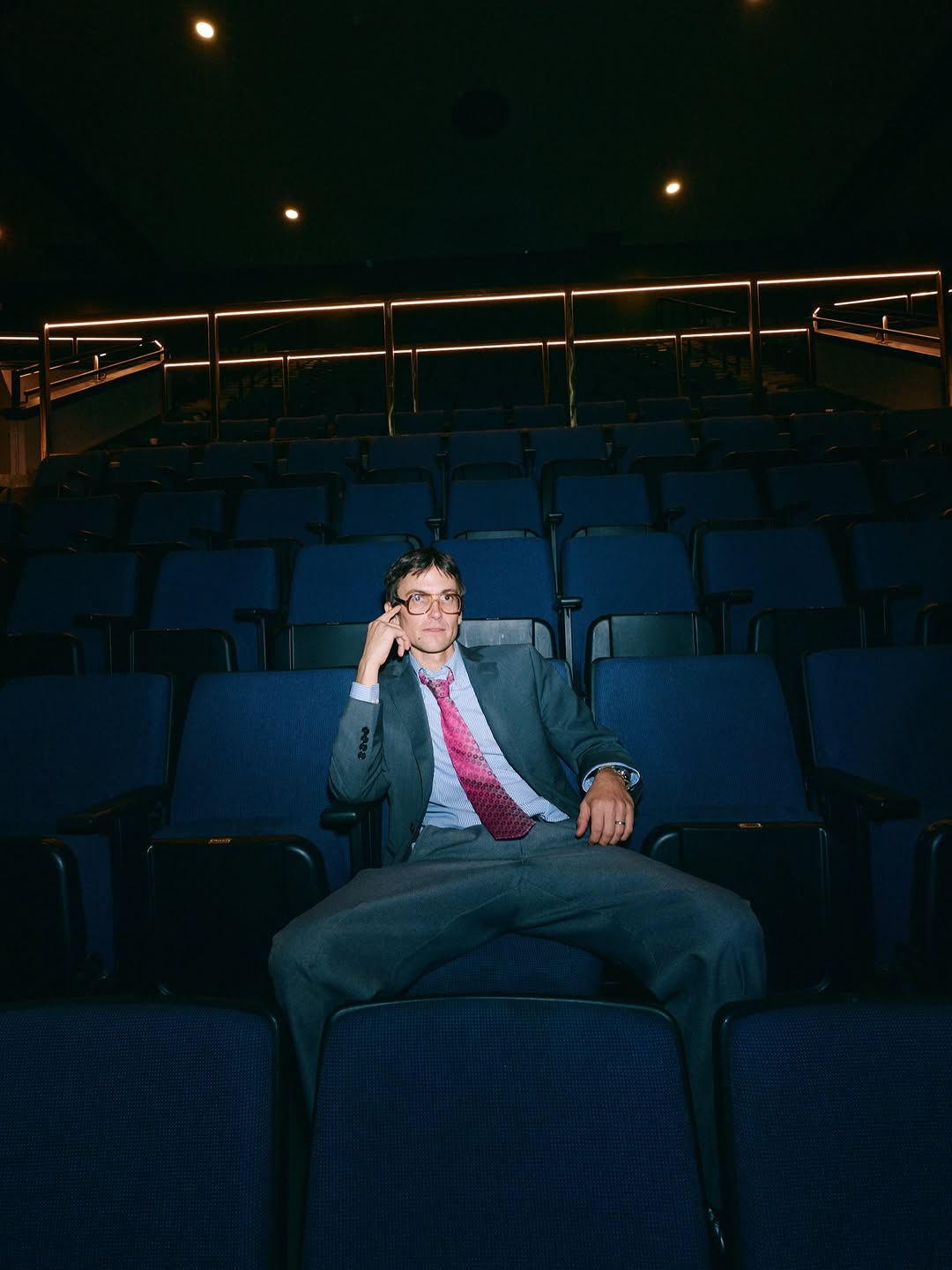 DIIV frontman Zachary Cole Smith sits alone in an empty theater wearing a gray suit and pink tie.