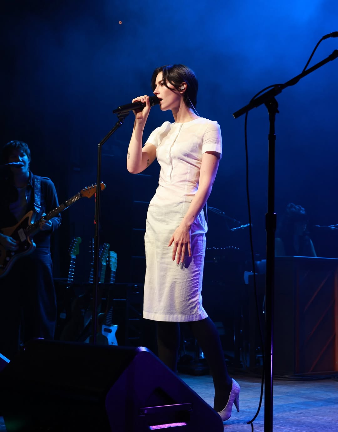 Gracie Abrams performs on stage in a white dress, singing into a microphone under blue lights.