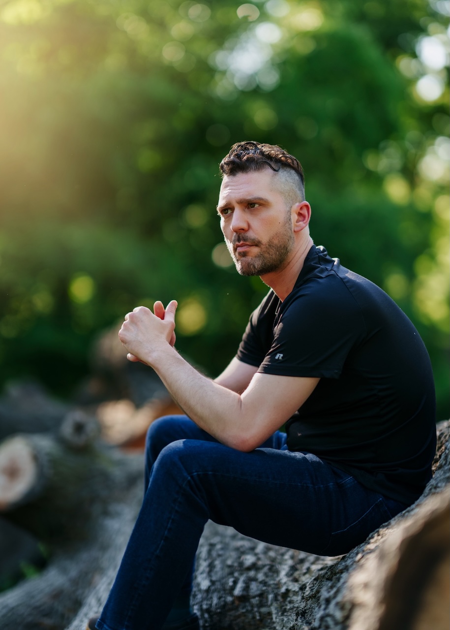 Joe Sparks seated outdoors on a log in a black shirt and jeans, hands clasped, looking at the camera