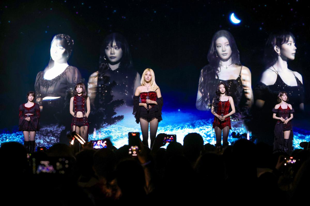 LE SSERAFIM perform on stage during their PUREFLOW tour, with all five members in red and black outfits before a starlit backdrop and the crowd in silhouette.