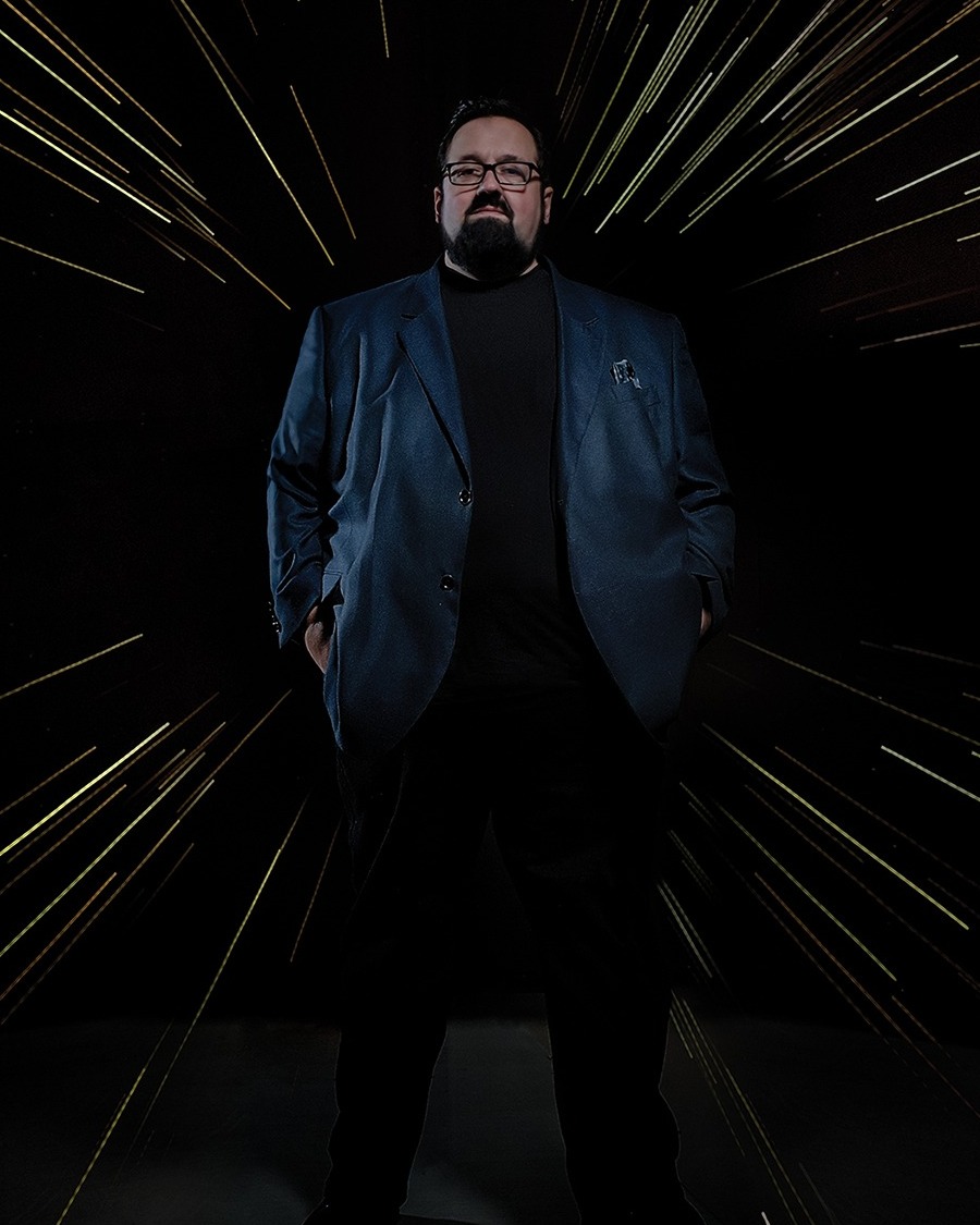 Christian McBride stands in a blue blazer and black turtleneck against a dark backdrop lit with streaks of light.