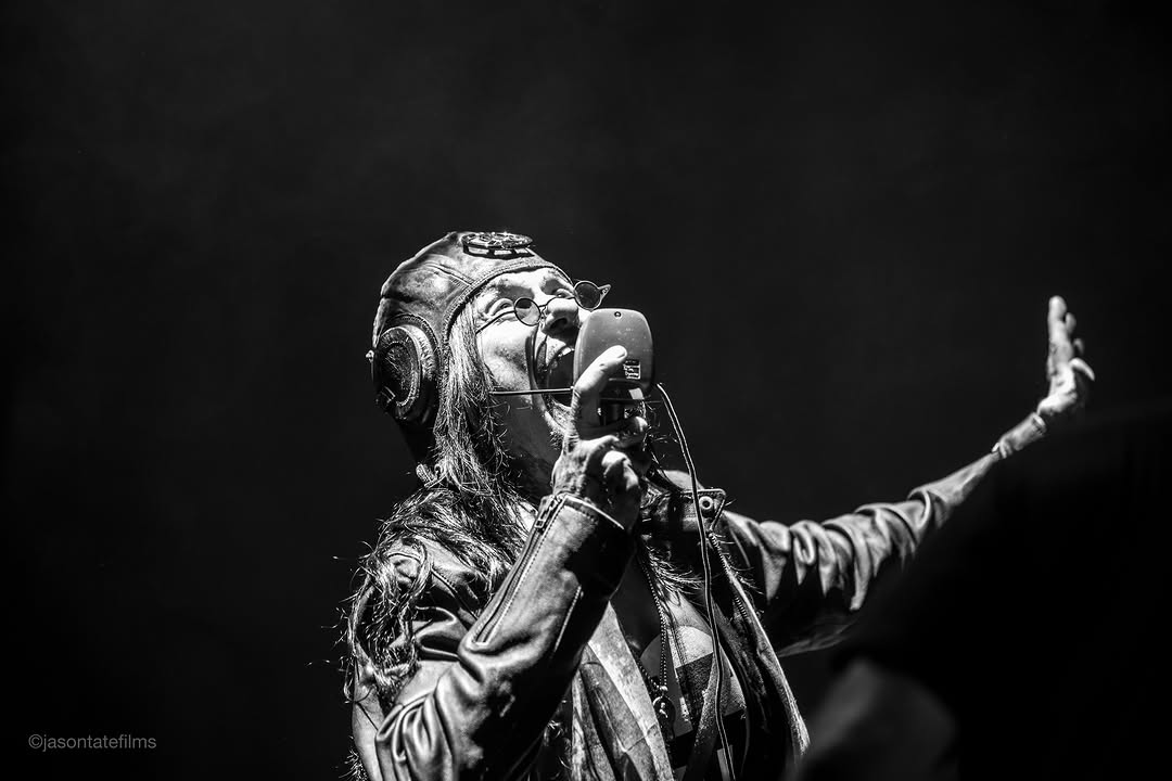 Al Jourgensen of Ministry performs live in a black and white photo, wearing an aviator cap and round glasses, singing into a microphone.