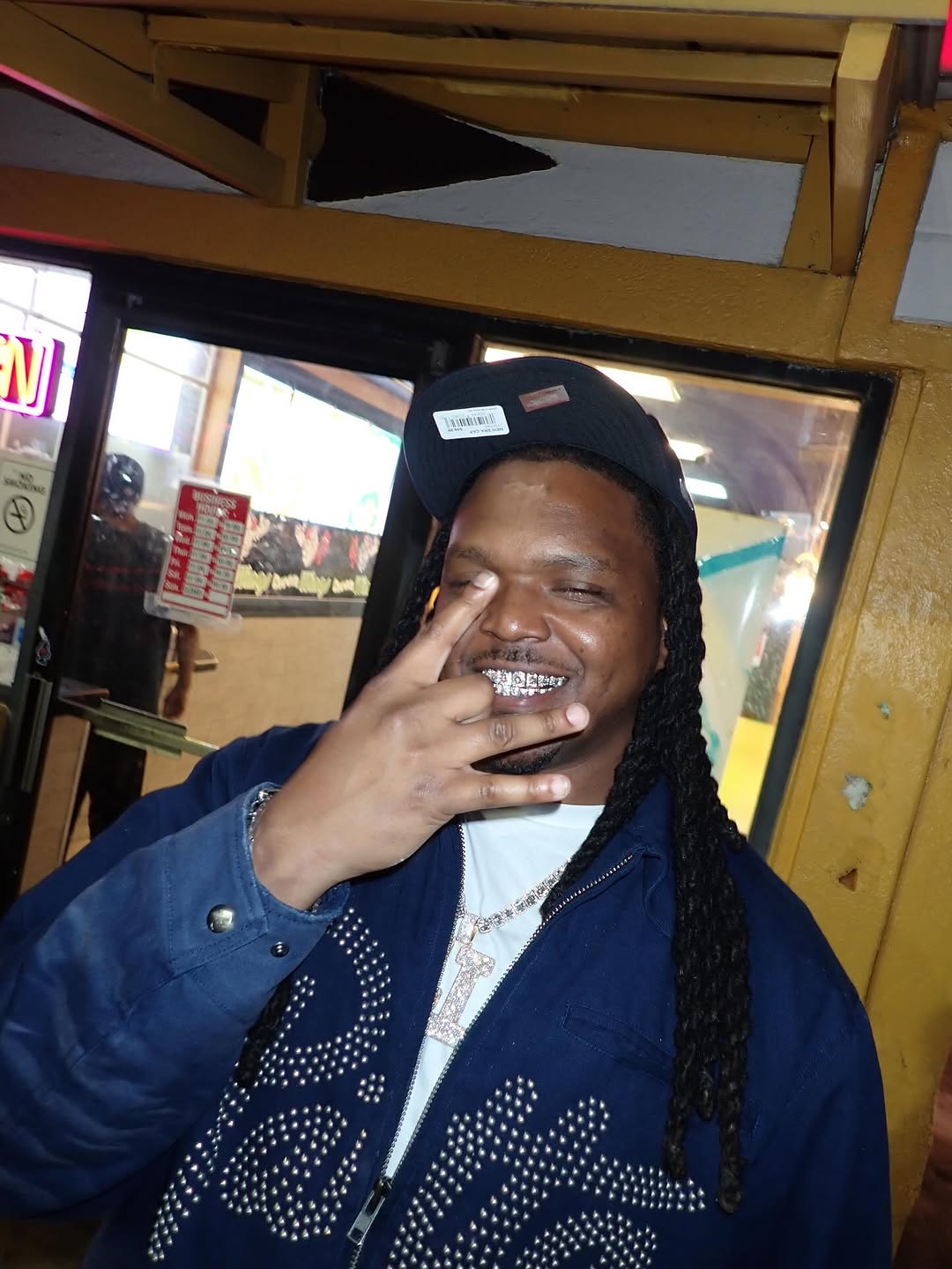 Young Nudy smiles in a blue studded jacket and black cap, showing a diamond grill.