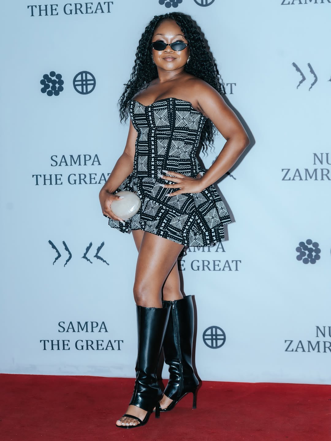 Sampa the Great poses on the red carpet at her Nu Zamrock album event in a black and white patterned dress and tall black boots.
