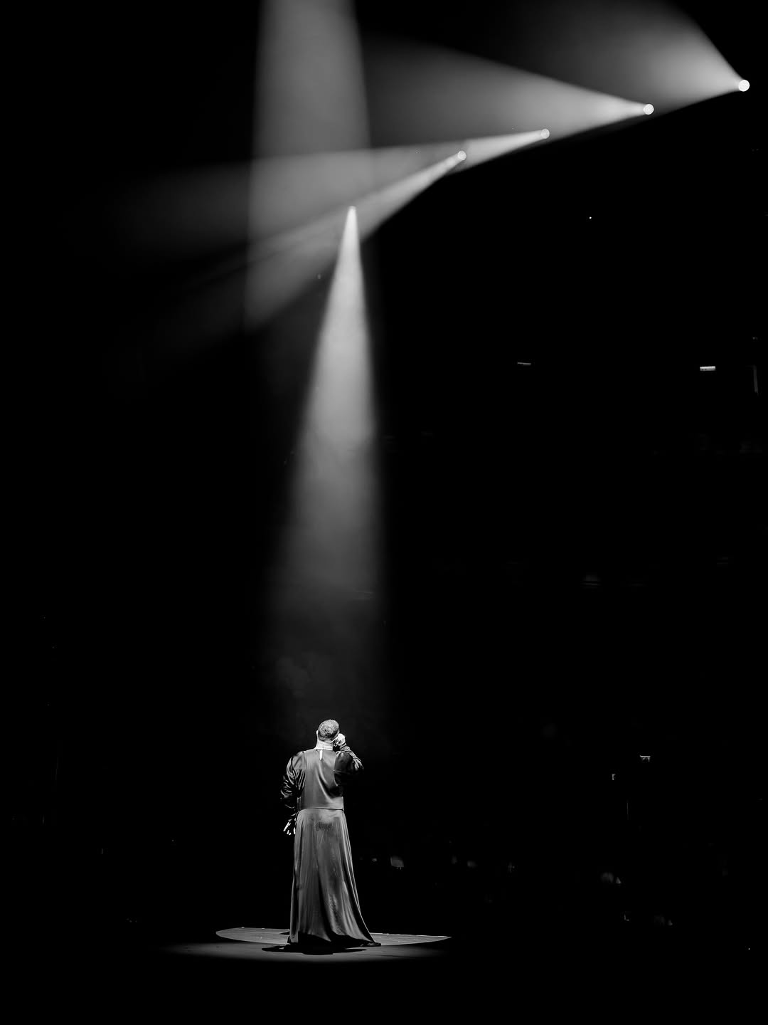 Sam Smith stands alone on a dark stage in a single spotlight, seen from behind, in black and white.