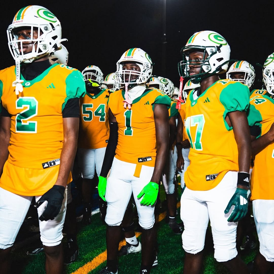 Miami Jackson Generals players in gold and green uniforms and white helmets stand together on the field during a night game
