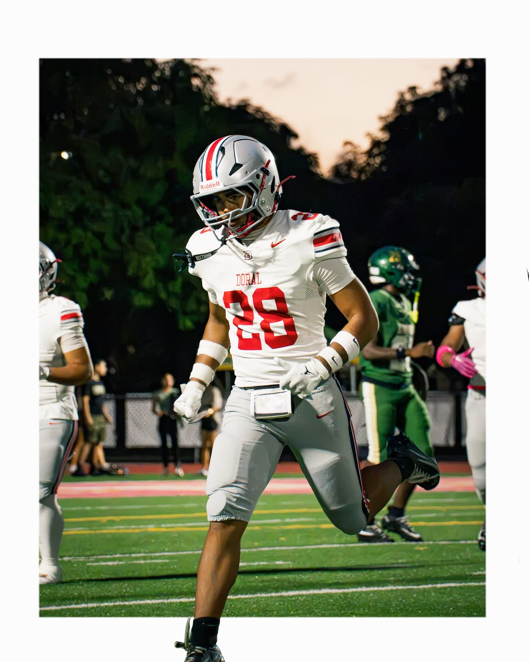 A Doral Academy Firebirds player in a white uniform with red trim, No. 28, runs on the field during a game at dusk