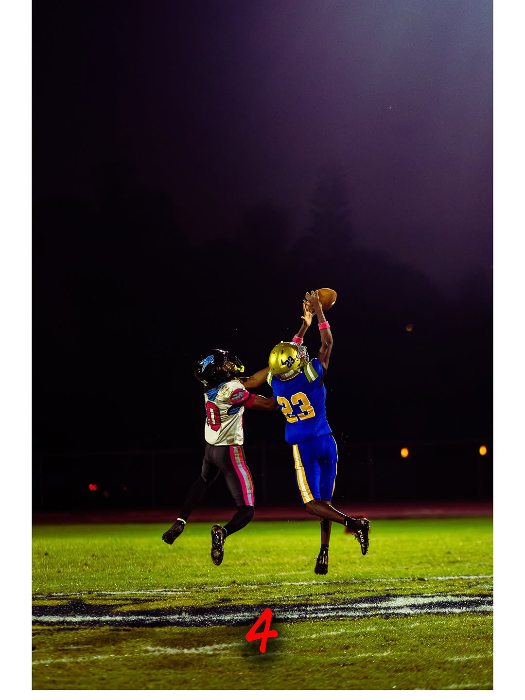 A West Broward Bobcats player in a blue and gold uniform leaps high to haul in a pass over a defender during a night game