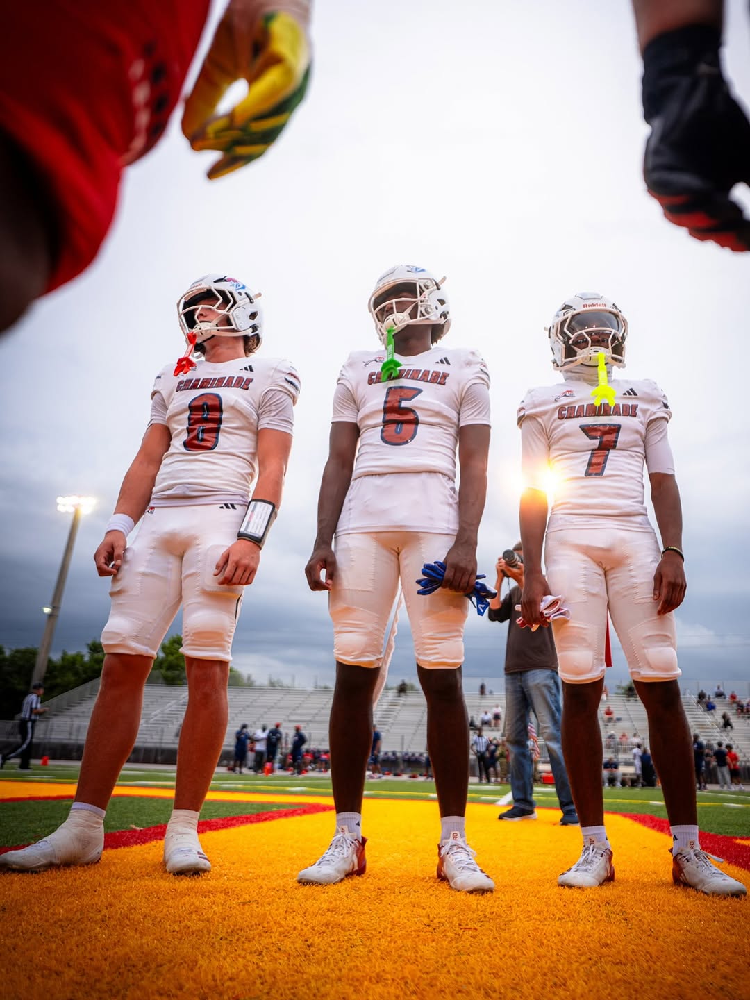 Three Chaminade-Madonna Lions players in white uniforms and helmets stand on the field before a game