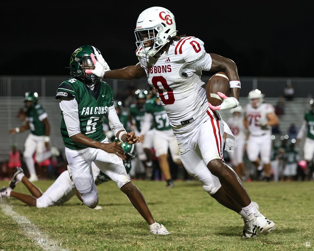 A Cardinal Gibbons Chiefs ball carrier in a white uniform stiff-arms a Flanagan defender while running with the ball during a night game