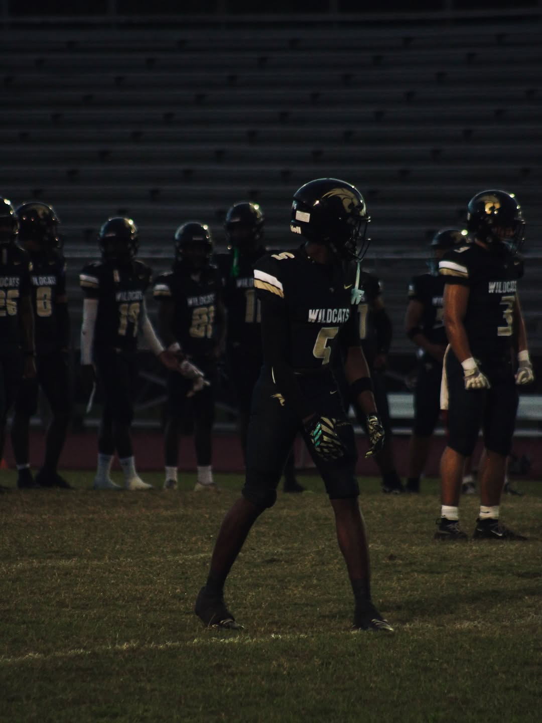 Western Wildcats players in black uniforms and helmets stand on the field during a night game in Davie, No. 5 in the foreground