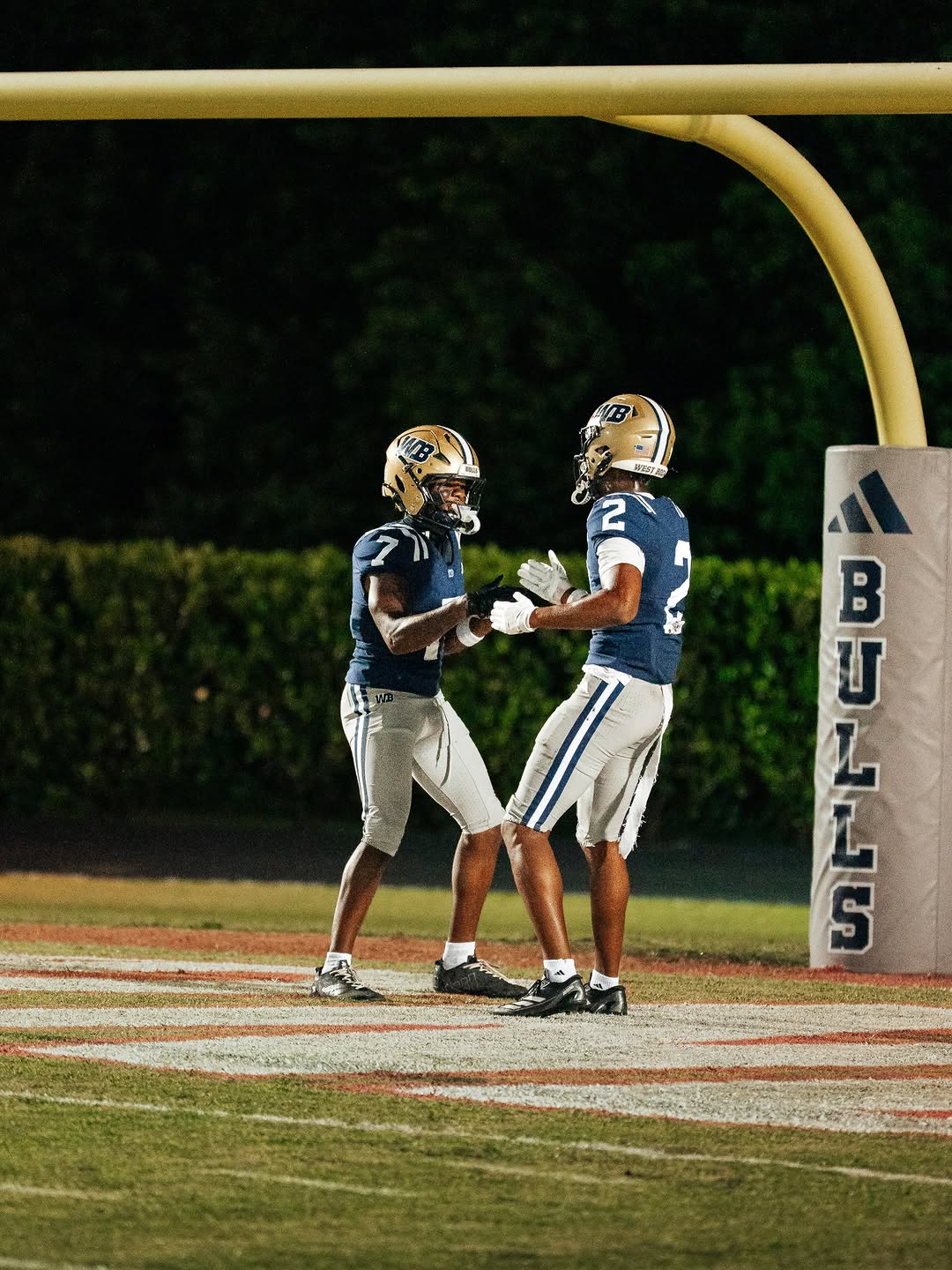Two West Boca Raton Bulls players in navy jerseys and gold helmets celebrate in the end zone under the goalpost during a night game