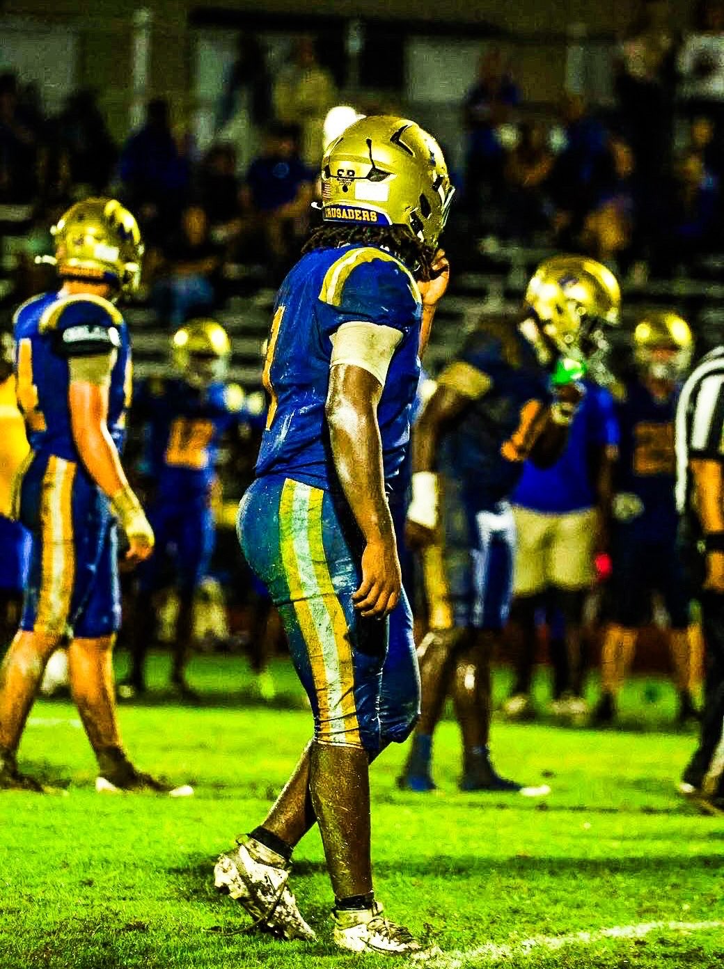 A Cardinal Newman Crusaders player in a muddy blue uniform and gold helmet walks on the field during a night game, teammates behind him