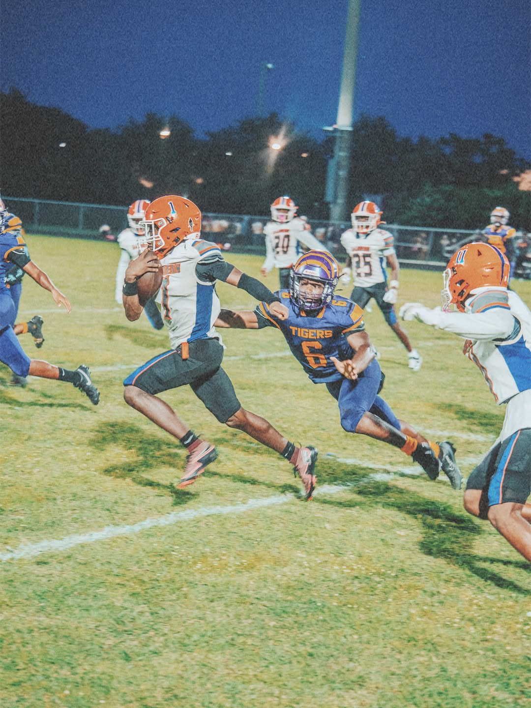 A Palm Beach Gardens Gators ball carrier in a white jersey and orange helmet runs with the ball as a defender reaches for him during a night game