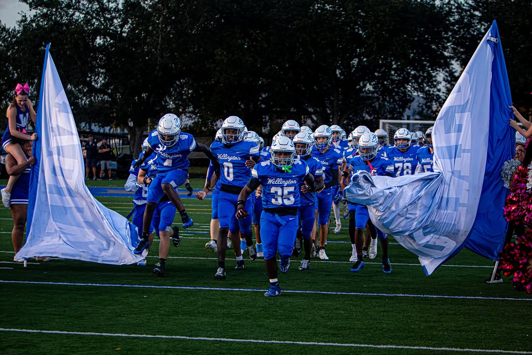 The Wellington Wolverines in blue uniforms and white helmets run onto the field through the team banner before a home game