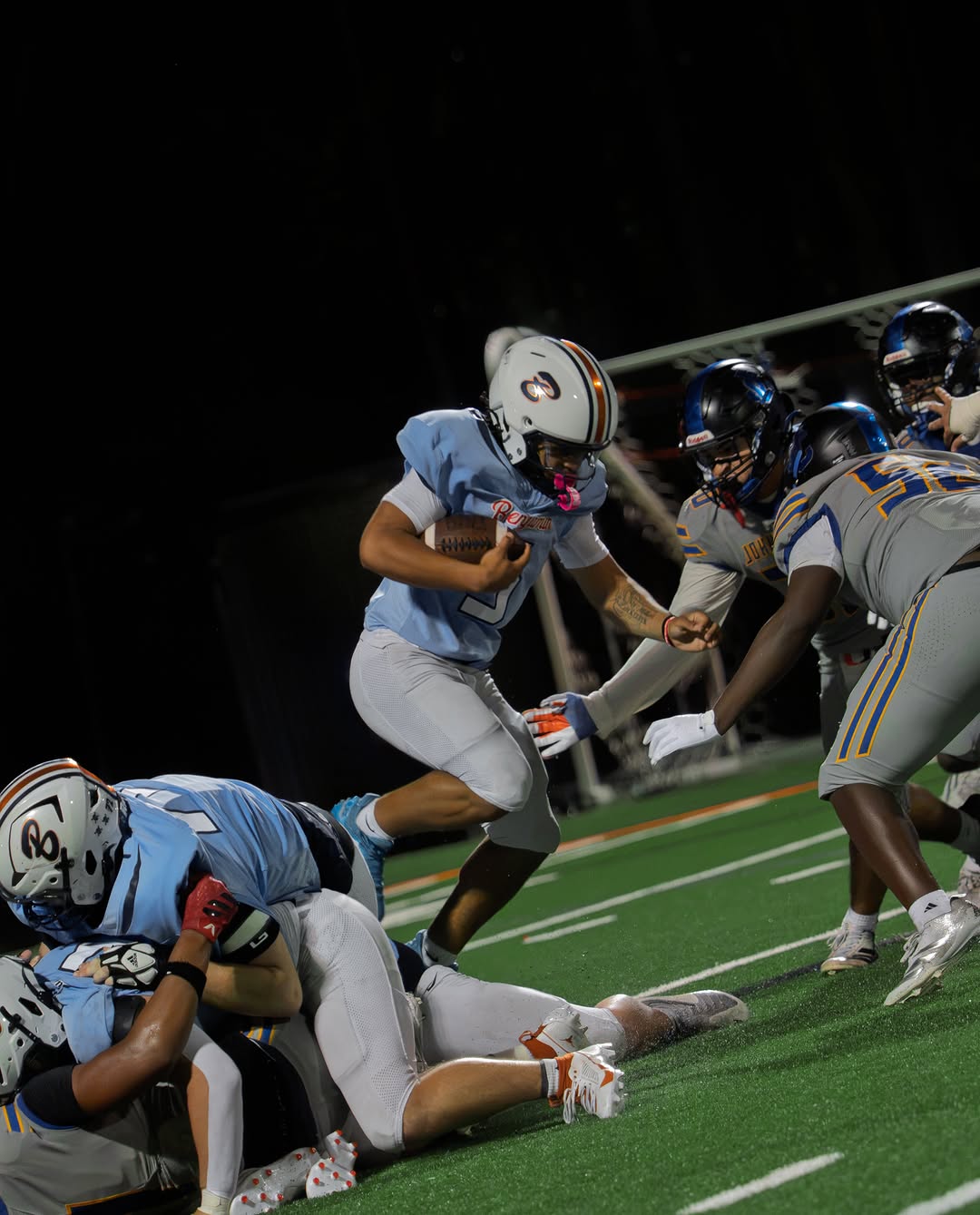 A Benjamin Buccaneers ball carrier in a light blue uniform breaks through the line with the ball as defenders close in during a night game