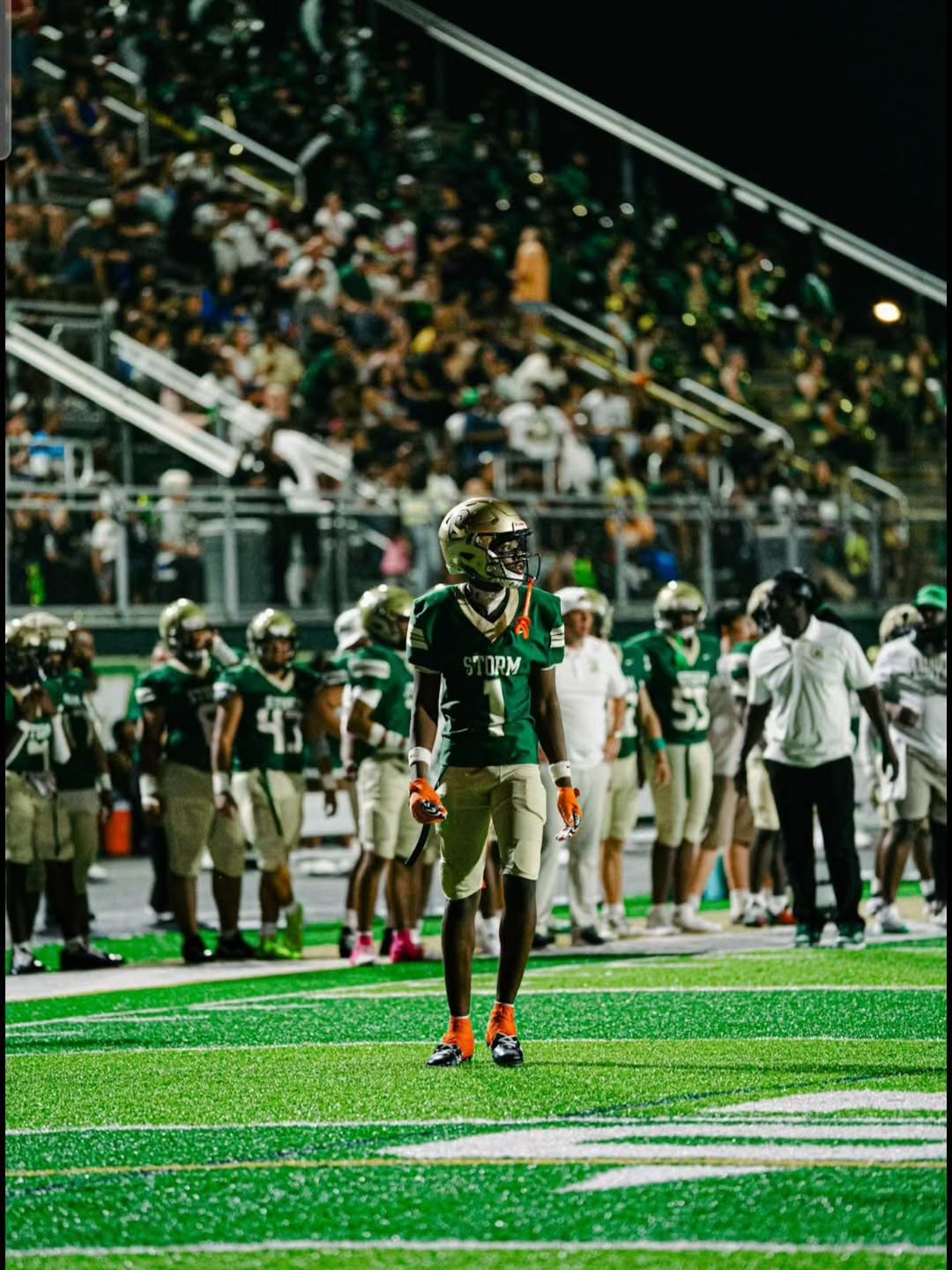Chamberlain Storm player in a green jersey and gold helmet standing on the field during a night game with teammates and fans behind him