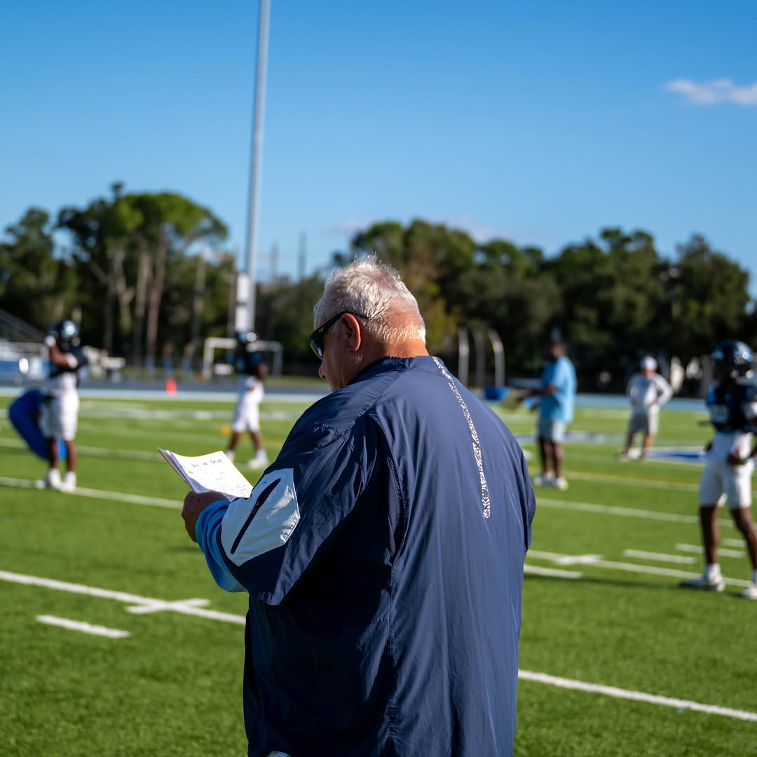 Berkeley Prep head coach watching over practice on the field, clipboard in hand, with Buccaneers players in helmets working behind him