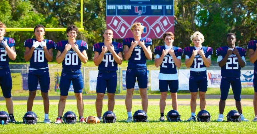 Palm Harbor University Hurricanes senior players in navy uniforms lined up on the field with their helmets at their feet in front of the scoreboard