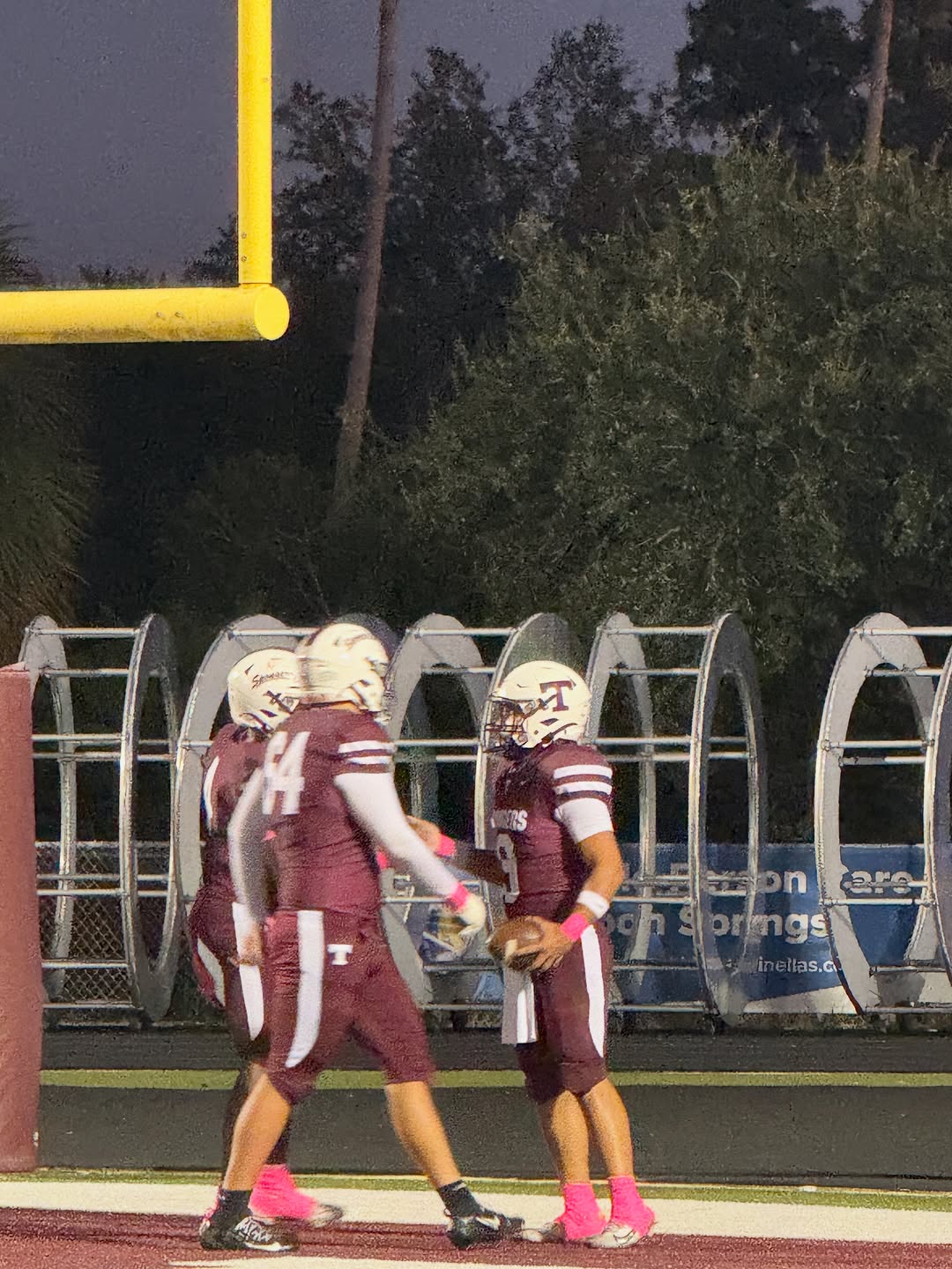 Tarpon Springs Spongers players in maroon uniforms celebrating in the end zone at night, one holding the football