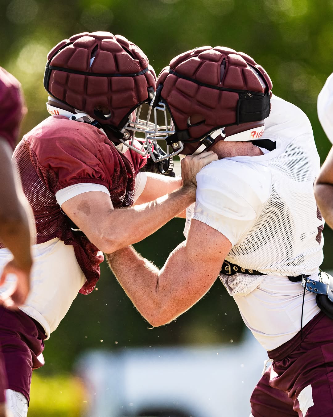 Two Riverview Rams players in maroon practice jerseys and padded helmet covers clash in a blocking drill at practice